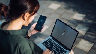 Young Asian businesswoman sitting on the bench in an urban park working outdoors, logging in to her laptop and holding smartphone on hand with a security key lock icon on the screen. Privacy protection, internet and mobile security concept