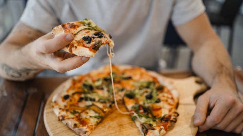 white young man with hat eating a pizza with vegetables on a wooden table