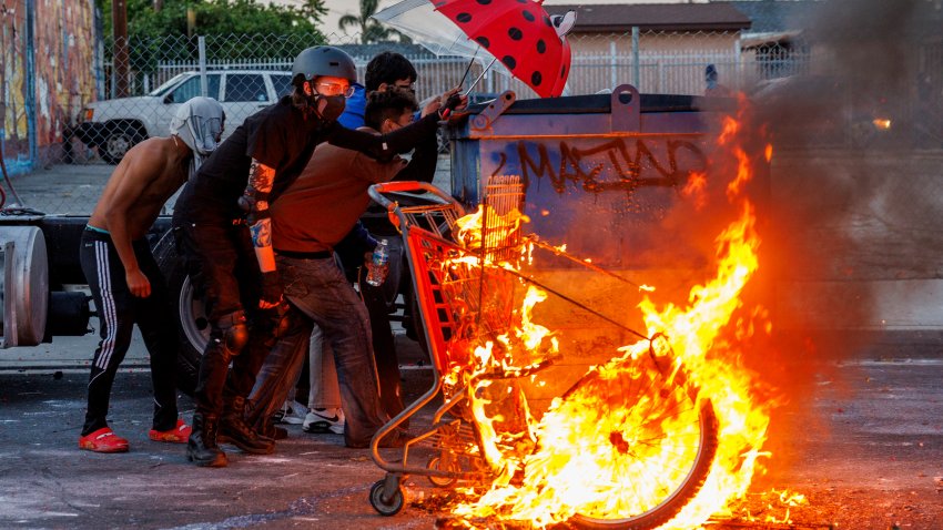 COMPTON, CA -JUNE 7, 2025: Protesters hide behind a dumpster near a set  fire as  LA County Sheriff deputies shoot projectiles to keep demonstrators from advancing  after ICE raids at a nearby Home Depot and the Garment District brought out resistance from Los Angeles residents on June 7, 2025 in Compton, California.(Gina Ferazzi / Los Angeles Times via Getty Images)
